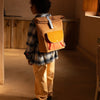 A person wearing a plaid jacket and cream pants stands in a warmly lit room, viewed from the back, showcasing a large Sticky Lemon Backpack | Farmhouse | Corduroy Harvest Moon.