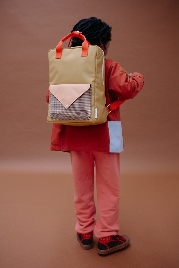 A person wearing red and pink clothes stands against a brown background, facing away, with the Sticky Lemon Backpack Large - Envelope in Bookworm Green—featuring orange straps and a front pocket with a triangular flap—crafted from recycled PET bottles for an eco-friendly touch.