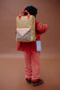 A person wearing red and pink clothes stands against a brown background, facing away, with the Sticky Lemon Backpack Large - Envelope in Bookworm Green—featuring orange straps and a front pocket with a triangular flap—crafted from recycled PET bottles for an eco-friendly touch.