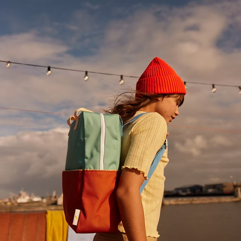 A young person in a red beanie and yellow shirt stands outdoors by water under string lights, carrying the Sticky Lemon Backpack Large in Aquatic Green + Shipwreck Rust, which features waterproof lining. Distant buildings and clouds are visible.