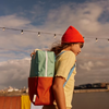 A young person in a red beanie and yellow shirt stands outdoors by water under string lights, carrying the Sticky Lemon Backpack Large in Aquatic Green + Shipwreck Rust, which features waterproof lining. Distant buildings and clouds are visible.