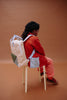 A child with long braided hair, wearing a red jacket and pink pants, sits on a white stool with the Sticky Lemon Backpack Large - Envelope - Twiggy Beige, made from recycled PET bottles, against a solid brown background.
