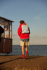 A person in a red hoodie, beige shorts, and red socks stands by a seaside kiosk facing the ocean, wearing a Sticky Lemon Backpack Large - Special Edition - Wonders of the Deep Sea, made from recycled PET bottles with blue anchor print lining.
