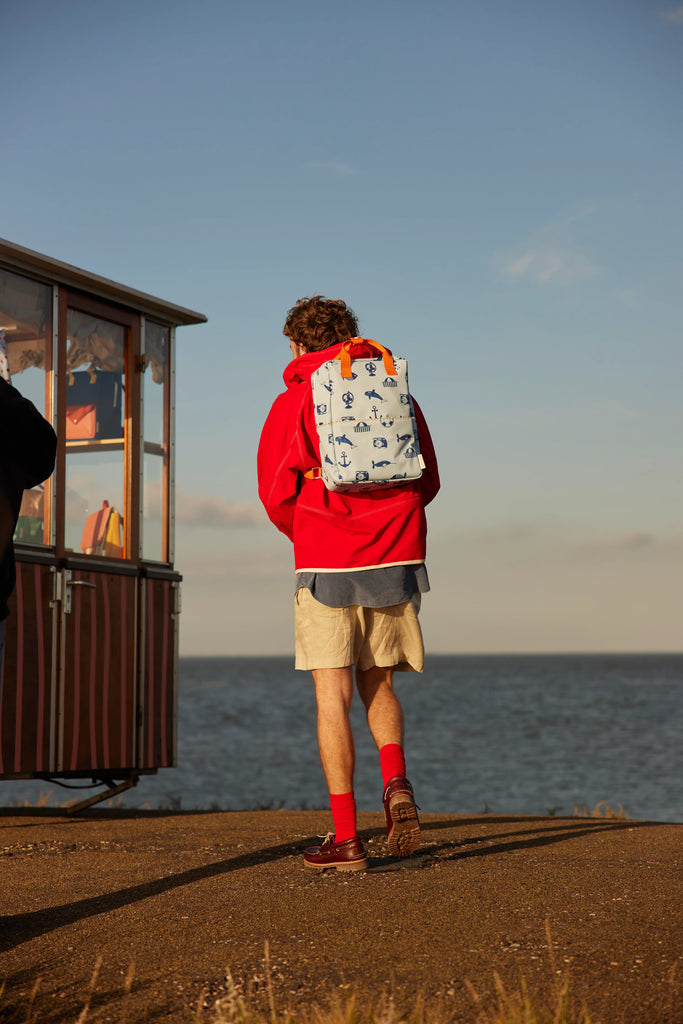 A person in a red hoodie, beige shorts, and red socks stands by a seaside kiosk facing the ocean, wearing a Sticky Lemon Backpack Large - Special Edition - Wonders of the Deep Sea, made from recycled PET bottles with blue anchor print lining.