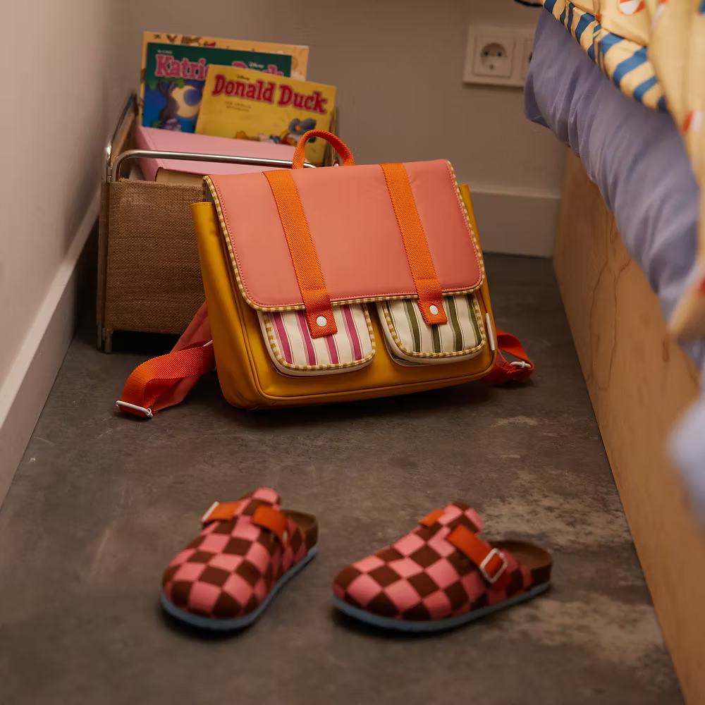 A Sticky Lemon School Bag | Coral Pink + Submarine Yellow, made from recycled PET and lined with waterproof nylon, sits by a box of children’s books and a Donald Duck comic; pink and brown checkered clogs rest near the bed.