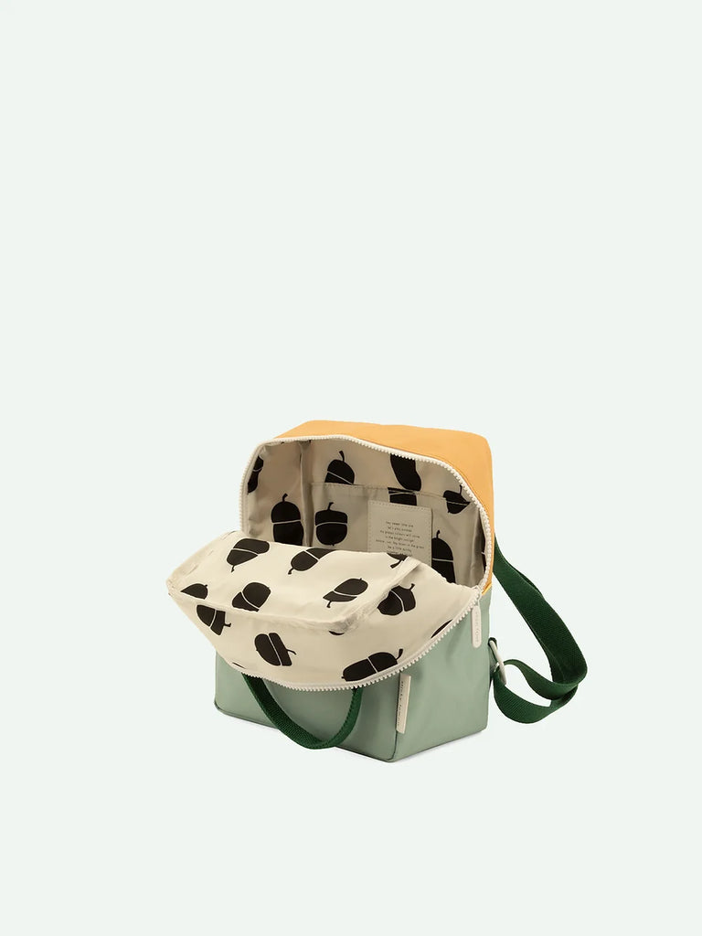 An open, stylish Sticky Lemon Backpack Small in the Meet Me In The Meadows color blocking design, featuring a green strap and a beige flap, showcasing a white interior patterned with black abstract shapes, against a pale background, and finished with a durable YKK zipper.