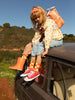 A young girl with a red headband sits on the roof of a car in a countryside setting, wearing colorful clothing and red sneakers, with a Sticky Lemon Backpack Small | Farmhouse | Special Edition resting on her shoulder.