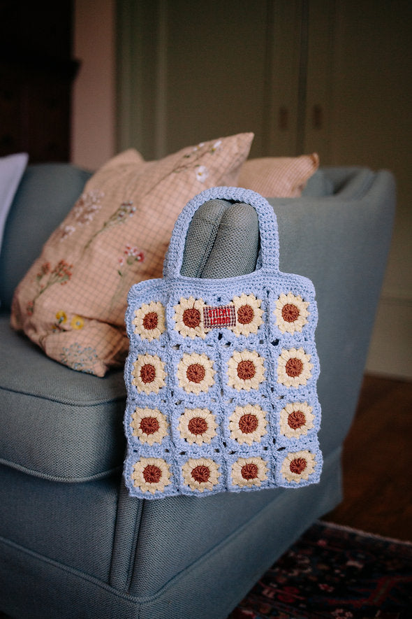 A Sticky Lemon Crochet Bag - Lemon Tree - Flower - Willow Basket, featuring blue, cream, and brown granny squares, hangs on the arm of a light blue sofa. Reminiscent of a charming Sticky Lemon kids bag, this crochet bag brings playful color to any space. A decorative pillow with floral embroidery sits in the background.
