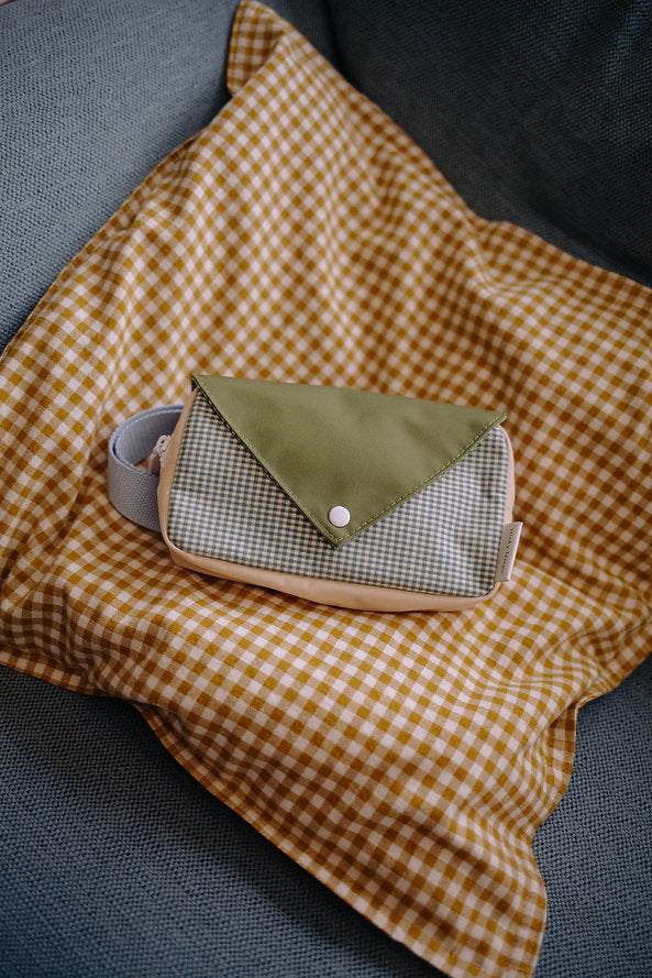 The Sticky Lemon Fanny Pack Small - Envelope - Tickling Grass, made from recycled PET bottles, features a green flap and checkered design. It sits atop a yellow and white gingham cloth on a gray couch.