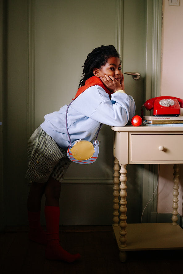 A child in a blue sweatshirt, red scarf, checkered shorts, and red socks leans on a beige table with a yellow Sticky Lemon Wallet Bag - Snail. On the table are a red rotary phone and stacked books.