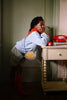 A child in a blue sweatshirt, red scarf, checkered shorts, and red socks leans on a beige table with a yellow Sticky Lemon Wallet Bag - Snail. On the table are a red rotary phone and stacked books.