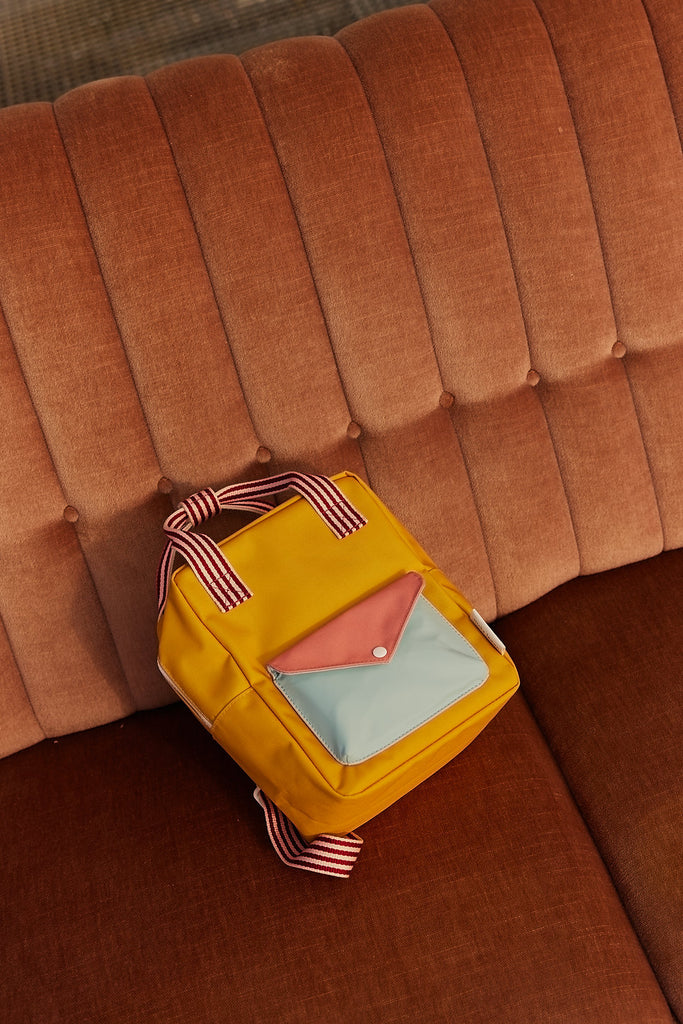The Sticky Lemon Backpack Small from the Envelope Collection in Submarine Yellow, featuring adjustable shoulder straps, a red and white striped handle, and a multicolored front pocket, sits on an orange tufted sofa.
