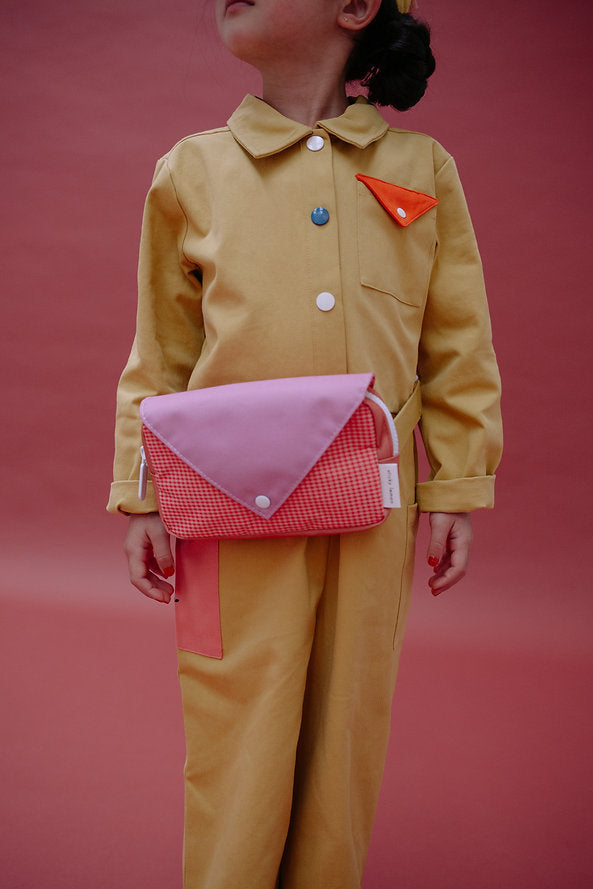 A child in a yellow jumpsuit with multicolored buttons stands against a pink background, wearing the Sticky Lemon Fanny Pack Small - Envelope - Rosebay Pink across their chest. The child's face is partially visible and looking upward.