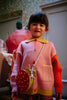 A young child with dark hair wears a pink and orange sweater, plaid pants, and the Sticky Lemon Wallet Bag - Ladybug—a round red bag with a yellow strap—standing indoors in a warmly lit room with floral wallpaper.