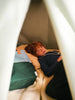 A young child with red hair, looking at the camera, is lying down inside a cozy blanket fort made with cushions and a Sticky Lemon Backpack Large in Meet Me In The Meadows color blocking pattern.