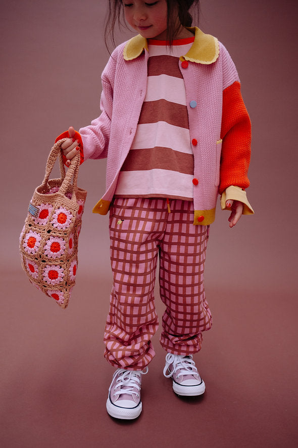 A child stands on a brown background wearing a colorful outfit with a pink and yellow jacket, striped shirt, plaid pants, white sneakers, and holding the charming Sticky Lemon Crochet Bag - Lemon Tree - Flower - Campfire Orange—a crochet bag adorned with cheerful flower patterns.