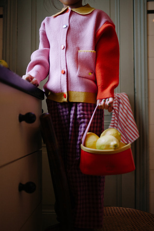 A child in a pink and red sweater and plaid pants stands indoors, holding a Sticky Lemon Treasure Bag - Campfire Orange filled with yellow pears. The child's face is not visible.
