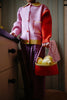 A child in a pink and red sweater and plaid pants stands indoors, holding a Sticky Lemon Treasure Bag - Campfire Orange filled with yellow pears. The child's face is not visible.
