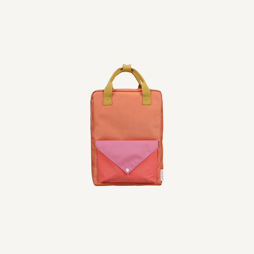 The Sticky Lemon Backpack Large - Envelope - Maple Whirl features a rectangular orange design with yellow handles, a front pocket with a pink flap and red checkered fabric, and adjustable shoulder straps, shown on a plain light background.