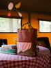 A stylish Sticky Lemon Backpack Large made from waterproof nylon is positioned upright on a red-and-white checkered tablecloth. Next to the backpack are books and a vase with yellow flowers. A cozy room with large windows serves