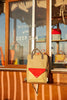 A Sticky Lemon Backpack Large - Envelope in Reed Green, featuring a beige body with a red front pocket and adjustable shoulder straps for comfort, hangs on a wooden counter in front of a window displaying "Wonders of the Deep Sea," with other colorful bags visible inside. The kids' bag is waterproof, making it both stylish and practical.