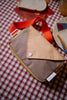 The Sticky Lemon Fanny Pack Small - Envelope - Apple Juice, featuring a brown and beige checkered design with an orange strap, sits on a red and white checkered tablecloth next to sliced bread and a plate.