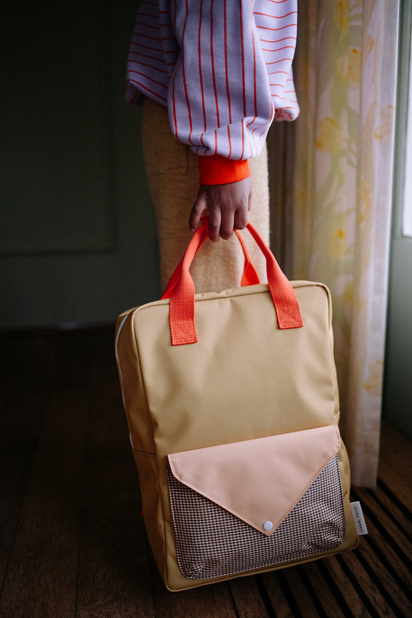 A person wearing a striped shirt holds the Sticky Lemon Backpack Large – Envelope in Bookworm Green, made from recycled PET bottles, featuring vibrant orange handles and a front pocket. The person stands indoors near a window with floral curtains.