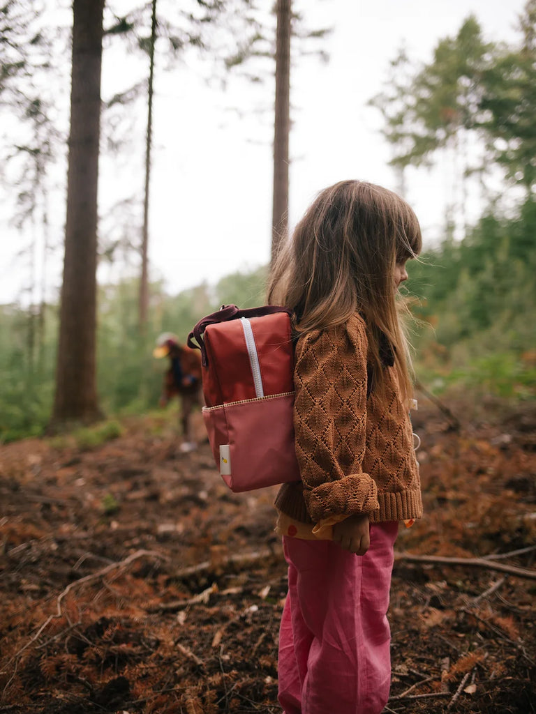 A young girl wearing a brown sweater and pink pants stands in a forest, carrying a Sticky Lemon Backpack Small in Love Story Red. She looks into the distance with tall trees in the background.