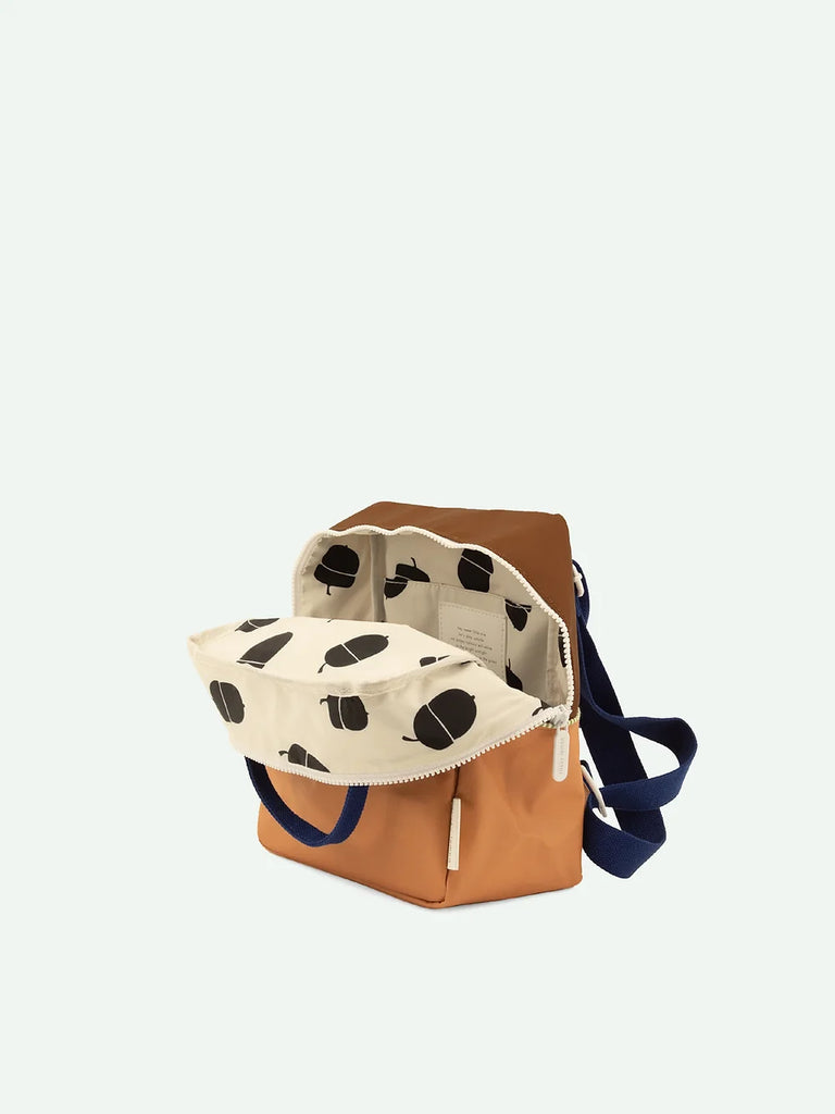A stylish, open Sticky Lemon Backpack Small in Treehouse Brown and white polka dots with a tan bottom and a navy blue strap, crafted from waterproof nylon against a plain light background.