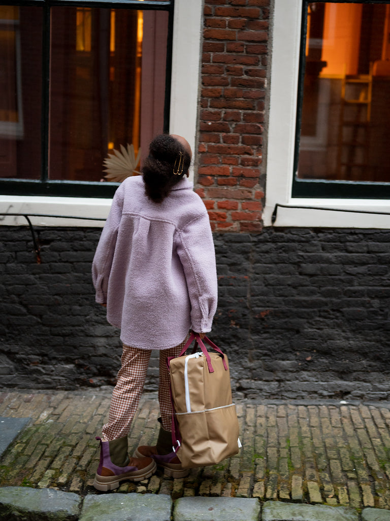 A woman standing on a cobblestone street, seen from behind, wearing a pink jacket and patterned pants, holding a Sticky Lemon Backpack | Uni | Inventor Green made of waterproof nylon. She looks toward a warmly lit window.