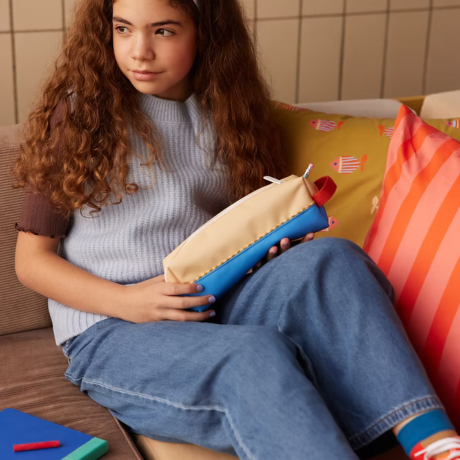 A young girl with long curly hair sits on a couch, holding the Sticky Lemon Pencil Case in Sandy Yellow + Radar Blue. She wears a light blue vest over a brown shirt and blue jeans, with colorful pillows and notebooks beside her.