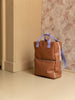 A Sticky Lemon Backpack | Uni | Buddy Brown with a YKK zipper and light purple straps stands against a corner with a beige and pink textured wall background.