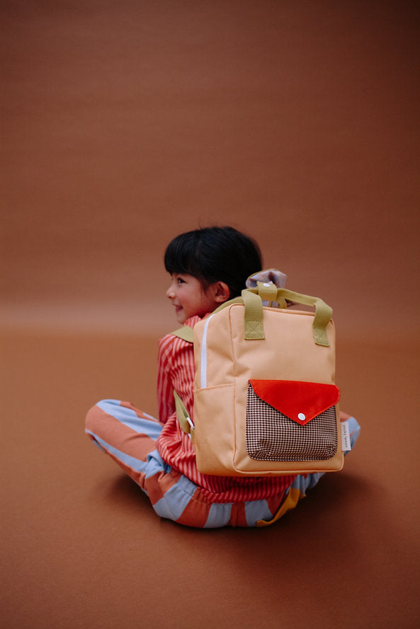 A young child with dark hair, smiling and wearing a striped colorful outfit, sits on the floor with a Sticky Lemon Backpack Small - Envelope - Apple Juice featuring a red and checkered front pocket. The background is solid brown.