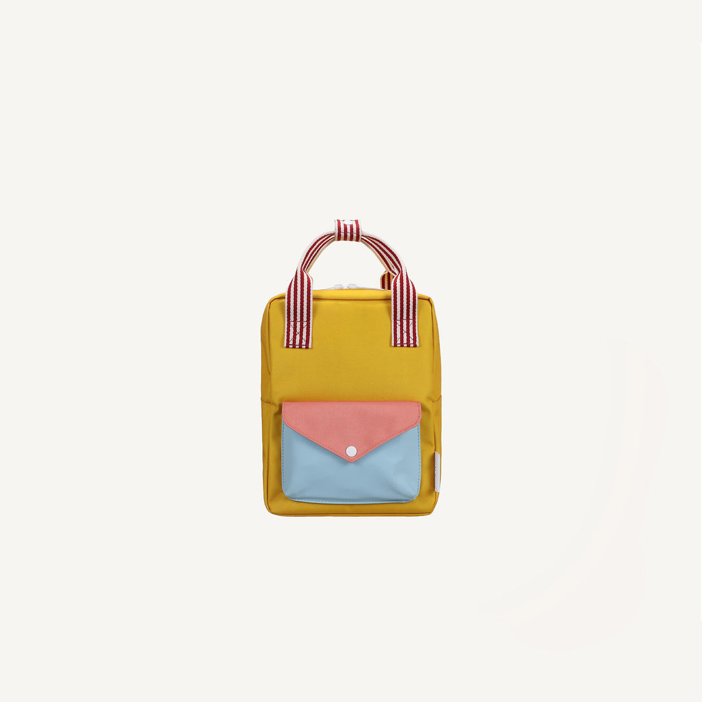 The Sticky Lemon Backpack Small from the Envelope Collection in Submarine Yellow features a blue and pink front pocket, white button clasp, adjustable shoulder straps, and red-and-white striped handles on a plain white background.