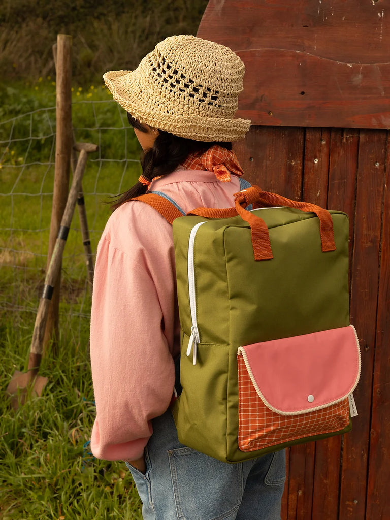 A woman viewed from behind, wearing a straw hat and a pink shirt, carries a Sticky Lemon Backpack Large in Sprout Green with an orange recycled RPET strap. She stands outdoors near a rustic wooden structure.