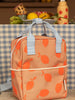 A peach-colored Sticky Lemon Backpack Small | Farmhouse | Special Edition with a white apple pattern, featuring a front pocket and blue handles, made from recycled PET bottles, stands on a wooden surface against a kitchen background with fruits.