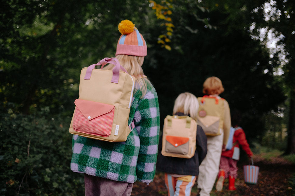 Four children with Sticky Lemon Backpack Medium - Envelope - Apple Grub walk through the woods. The first child wears a green checkered jacket and a colorful hat with a yellow pom-pom. These backpacks have waterproof lining—ideal for forest adventures.