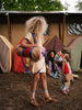 A girl with curly blonde hair stands at a campsite, backpack on, facing a canvas tent. Another child with a Sticky Lemon Duffle Bag from the Adventure Collection in Cousin Clay, and a seated woman can be seen in the background.