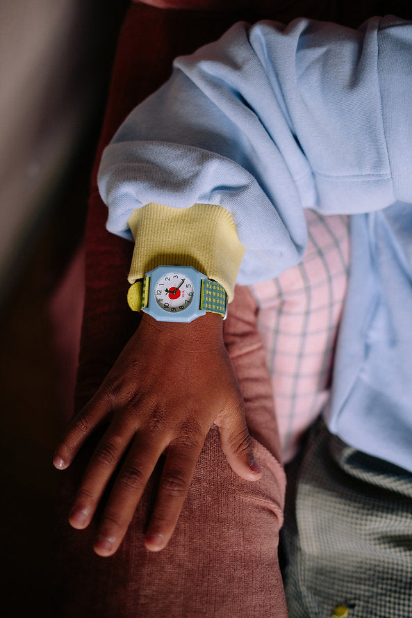A child in a blue sweatshirt with yellow sleeves wears the Sticky Lemon x Mini Kyomo Watch - Apple, featuring a colorful cartoon face and flower print strap, while resting their hand on a plaid cushion.