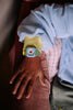 A child in a blue sweatshirt with yellow sleeves wears the Sticky Lemon x Mini Kyomo Watch - Apple, featuring a colorful cartoon face and flower print strap, while resting their hand on a plaid cushion.