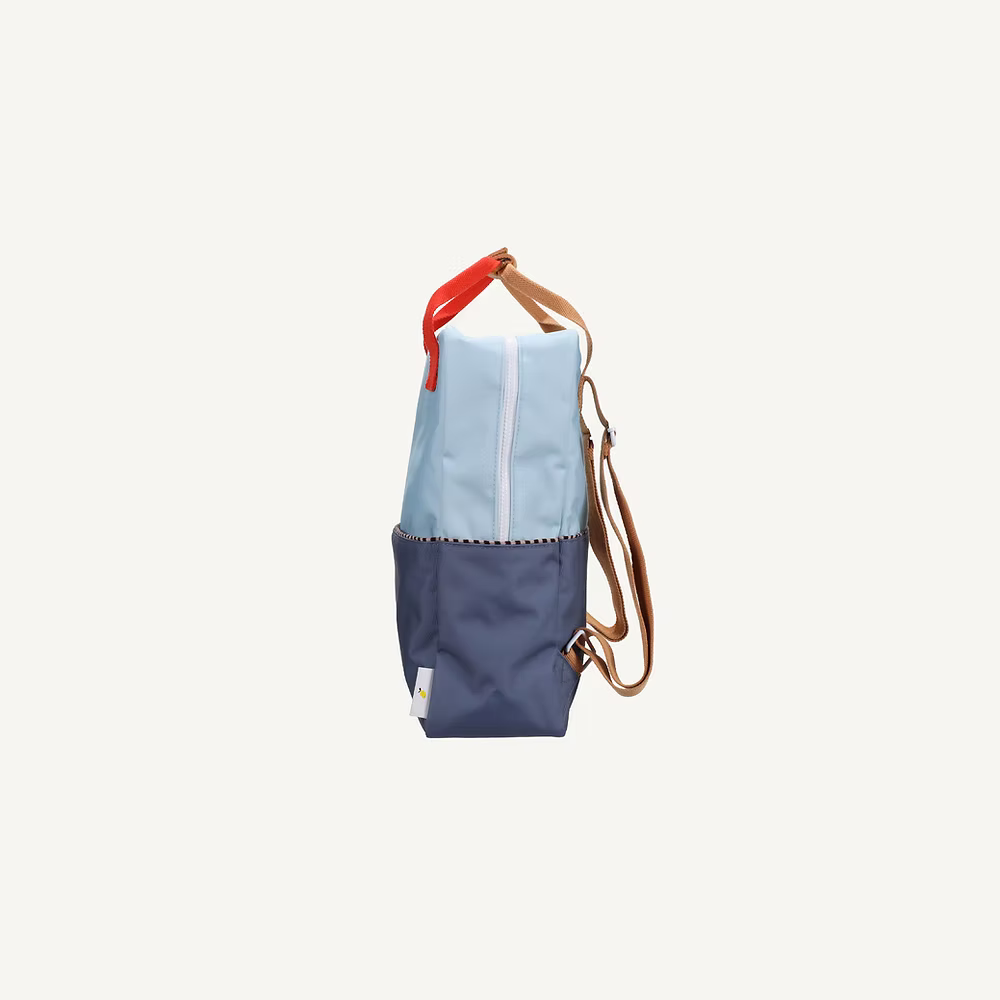 Side view of the Sticky Lemon Backpack Large | Color Blocking | Bissou Blue + Blue Whale, made from recycled PET bottles with a light blue top, dark blue bottom, tan straps, red handle, and front pocket against a white background.