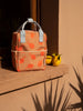 A peach-colored Sticky Lemon backpack Small | Farmhouse | Special Edition with a red apple print, crafted from recycled PET bottles, positioned against a sunlit beige wall next to two lemons, casting sharp shadows on a warm, sunny day.