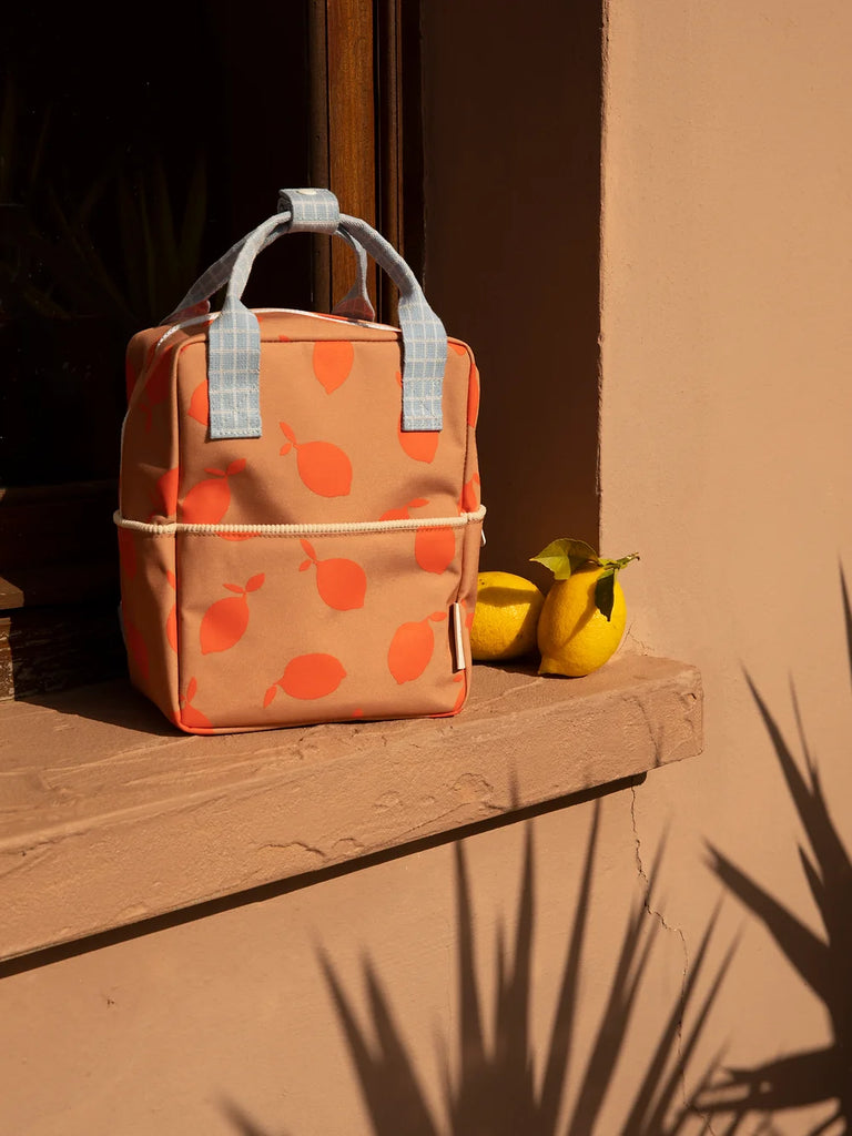 A peach-colored Sticky Lemon backpack Small | Farmhouse | Special Edition with a red apple print, crafted from recycled PET bottles, positioned against a sunlit beige wall next to two lemons, casting sharp shadows on a warm, sunny day.