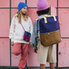 Two children stand in front of pink lockers. One child, wearing a blue beanie and white hoodie, leans against the lockers with a small purple bag featuring waterproof lining. The other child, in a pink beanie and gray shirt, faces away, showcasing an impressive Sticky Lemon Backpack Large | Color Blocking | Skate Ramp + Golden Cup with adjustable shoulder straps made from recycled PET.