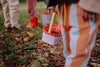 Children holding Sticky Lemon Treasure Bags in Dewy Lilac, standing on grassy, leafy ground. Focus is on the lilac bag and striped pants, with other children carrying their own treasure bags in the background.