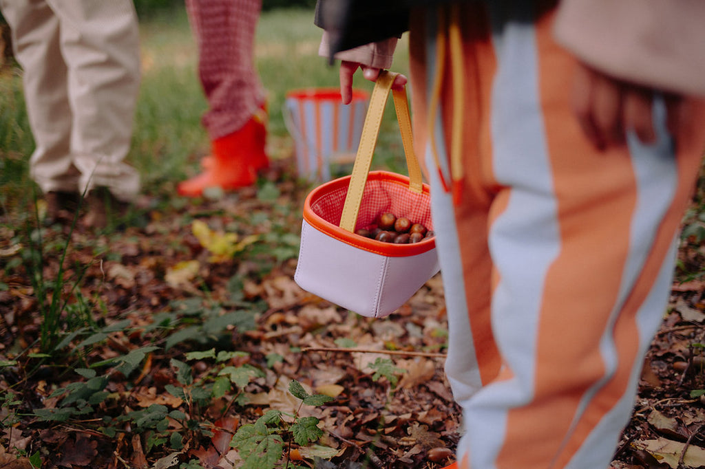 Children holding Sticky Lemon Treasure Bags in Dewy Lilac, standing on grassy, leafy ground. Focus is on the lilac bag and striped pants, with other children carrying their own treasure bags in the background.
