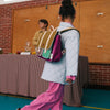 A young child with a Sticky Lemon School Bag in Gymnastic Pink featuring padded shoulder straps walks past a table draped with a striped cloth. A person sits behind the table, and the room, showcasing brick walls and a wooden cabinet, presents an intriguing backdrop. The child is dressed in a light blue quilted jacket and pink pants.