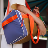 A person with a light-colored shirt holds the Sticky Lemon Sports Bag | Better Together—color-blocked in purple, brown, and orange with bright orange handles—made from recycled PET bottles and featuring a waterproof lining.
