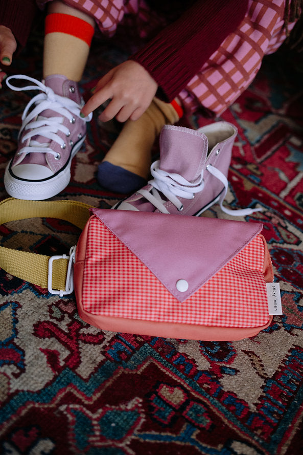 A child in pink checkered pants and mismatched socks ties light purple sneakers. In the foreground, a Sticky Lemon Fanny Pack Small - Envelope - Rosebay Pink rests on a patterned rug.