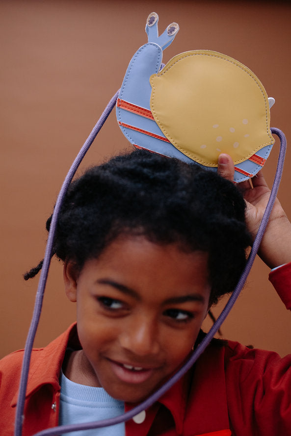 A dark-haired child smiles while holding the Sticky Lemon Wallet Bag - Snail on their head, dressed in a red jacket and light blue shirt, set against a plain brown background.